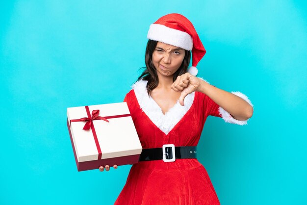 Young hispanic woman dressed as mama noel holding a gift isolated on blue background showing thumb down with negative expression