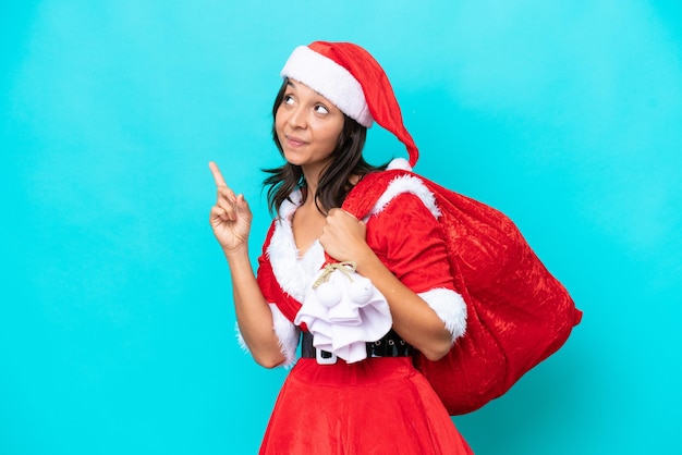 Young hispanic woman dressed as mama noel holding a sack isolated on blue background pointing up a great idea