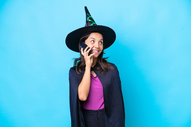 Young hispanic woman dressed as witch over isolated background keeping a conversation with the mobile phone