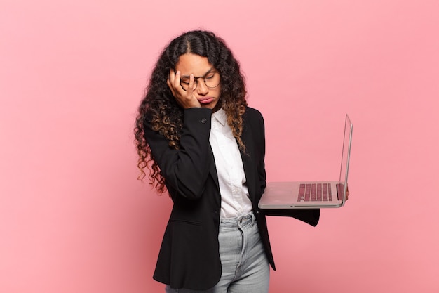 Young hispanic woman feeling bored, frustrated and sleepy after a tiresome, dull and tedious task, holding face with hand. laptop concept