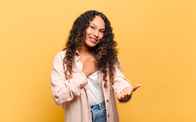 Young hispanic woman feeling happy and in love, smiling with one hand next to heart and the other stretched up front