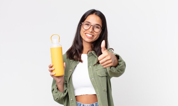 Young hispanic woman feeling proud,smiling positively with thumbs up and holding a coffee thermos