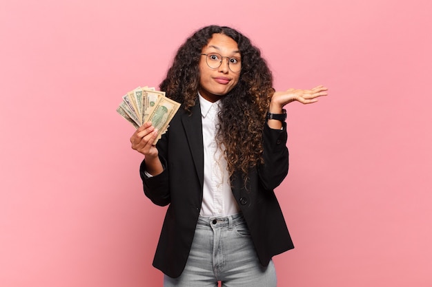 Young hispanic woman feeling puzzled and confused, doubting, weighting or choosing different options with funny expression. dollar banknotes concept