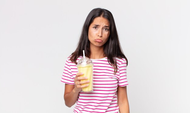 Young hispanic woman feeling sad and whiney with an unhappy look and crying and holding a vanila smoothy milkshake