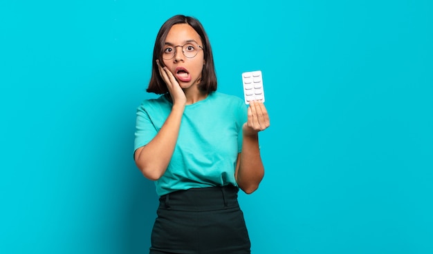 Young hispanic woman feeling shocked and scared, looking terrified with open mouth and hands on cheeks