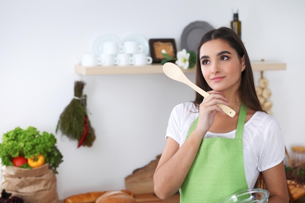 Young Hispanic woman in a green apron cooking in the kitchen Housewife holding wooden spoon while smiling