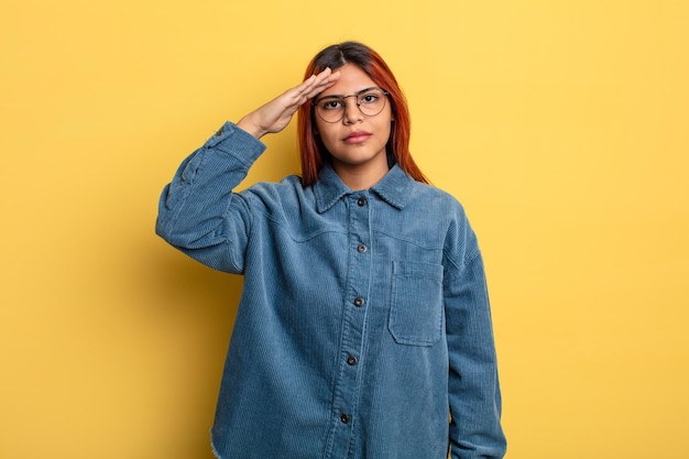 Young hispanic woman greeting the camera with a military salute in an act of honor and patriotism, showing respect