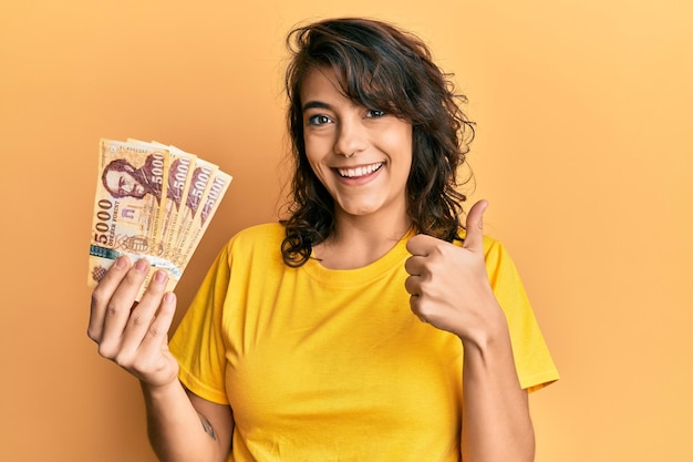 Young hispanic woman holding 5000 hungarian forint banknotes smiling happy and positive thumb up doing excellent and approval sign