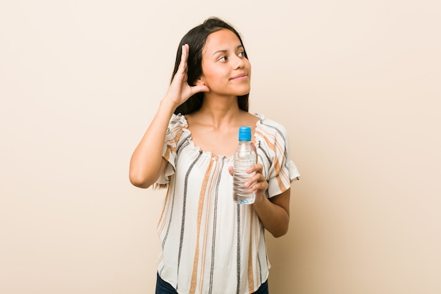 Young hispanic woman holding a bottle of water trying to listening a gossip.