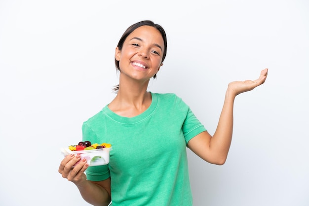 Young hispanic woman holding a bowl of fruit isolated on white background extending hands to the side for inviting to come