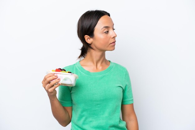 Young hispanic woman holding a bowl of fruit isolated on white background looking to the side