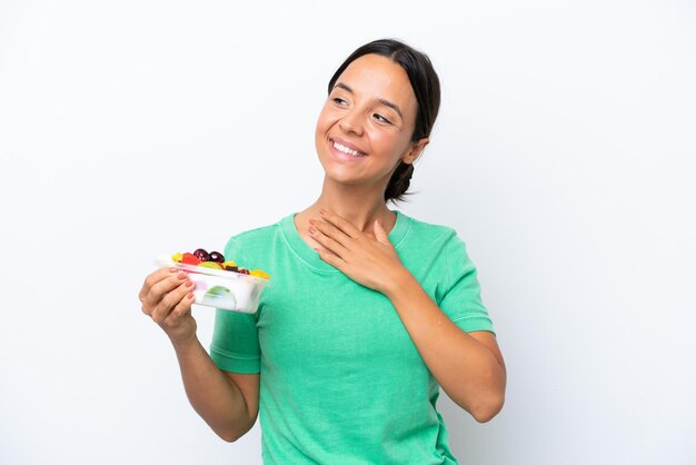 Young hispanic woman holding a bowl of fruit isolated on white background looking up while smiling