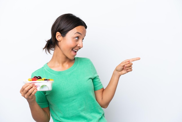 Young hispanic woman holding a bowl of fruit isolated on white background pointing finger to the side and presenting a product