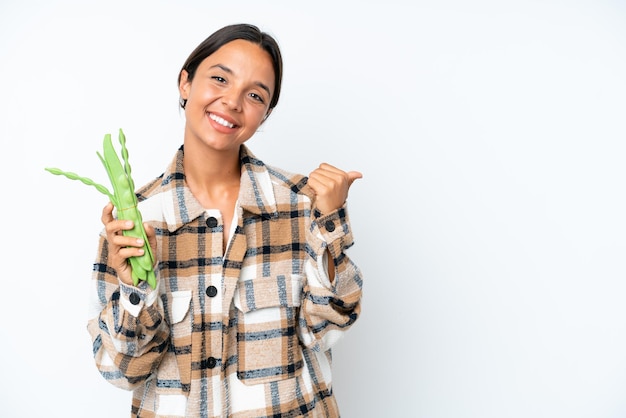 Young hispanic woman holding a green beans isolated on white background pointing to the side to present a product