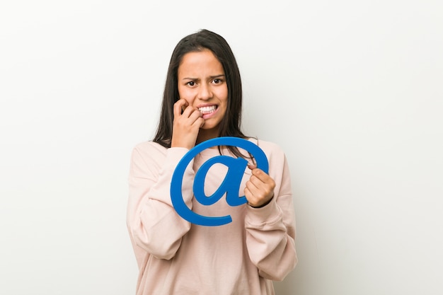 Young hispanic woman holding an at icon biting fingernails, nervous and very anxious.