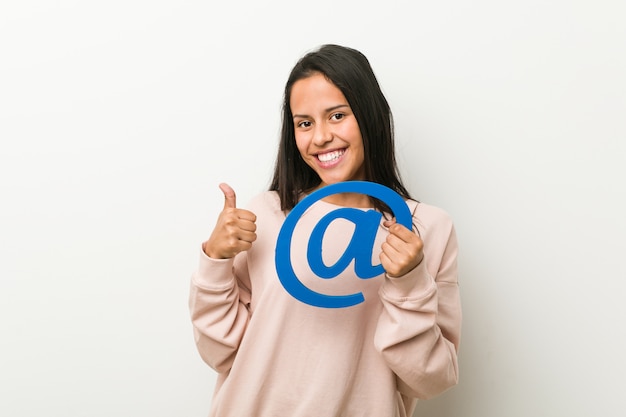 Young hispanic woman holding an at icon smiling and raising thumb up