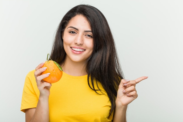 Young hispanic woman holding an orange smiling cheerfully pointing with forefinger away.