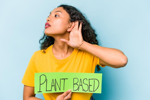 Young hispanic woman holding plant based placard isolated on blue background trying to listening a gossip