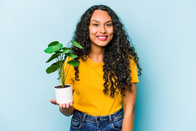 Young hispanic woman holding a plant isolated on blue background happy smiling and cheerful