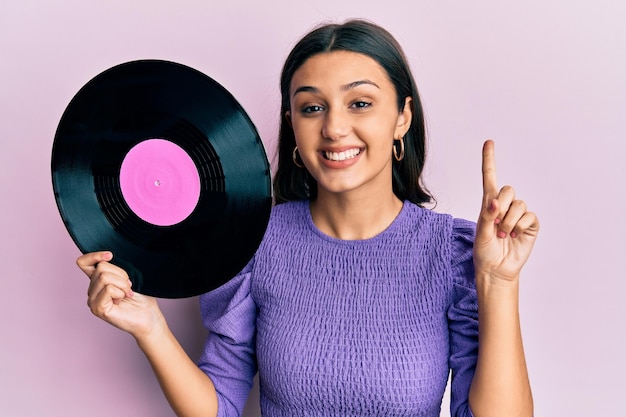 Young hispanic woman holding vinyl disc smiling with an idea or question pointing finger with happy face number one