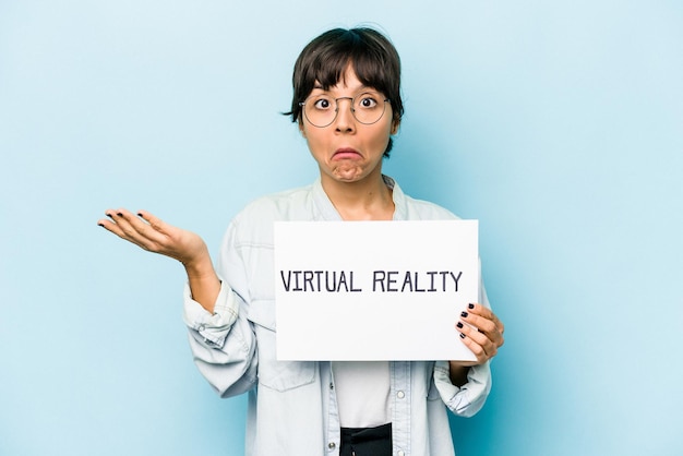 Young hispanic woman holding a virtual reality placard isolated on blue background shrugs shoulders and open eyes confused