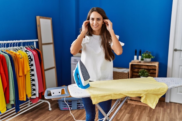 Young hispanic woman ironing clothes at laundry room covering ears with fingers with annoyed expression for the noise of loud music. deaf concept.