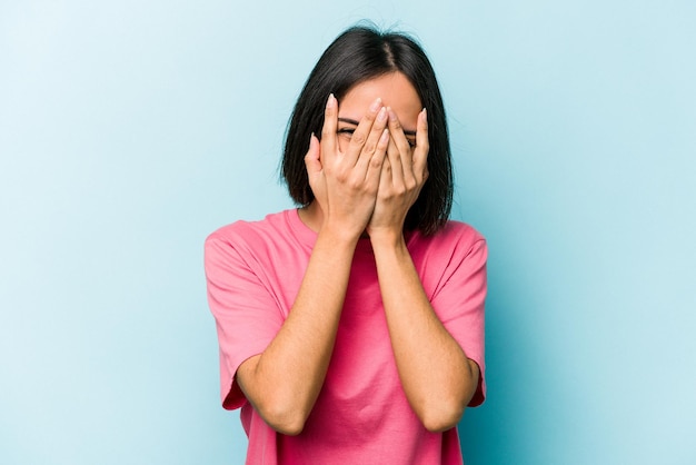 Young hispanic woman isolated on blue background blink at the camera through fingers embarrassed covering face