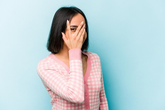 Young hispanic woman isolated on blue background blink at the camera through fingers embarrassed covering face