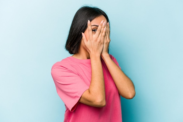 Young hispanic woman isolated on blue background blink through fingers frightened and nervous