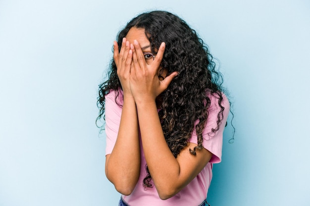 Young hispanic woman isolated on blue background blink through fingers frightened and nervous