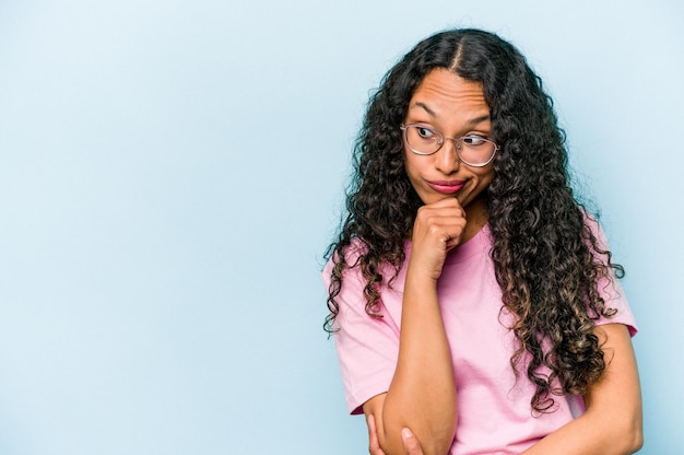 Young hispanic woman isolated on blue background looking sideways with doubtful and skeptical expression