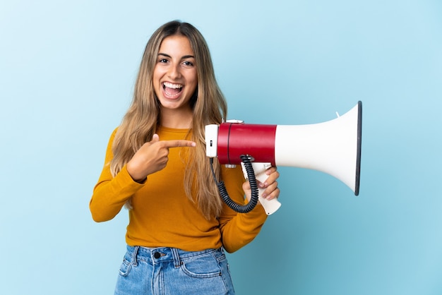 Young hispanic woman over isolated blue wall holding a megaphone and pointing side