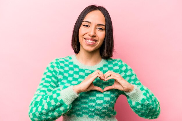 Young hispanic woman isolated on pink background smiling and showing a heart shape with hands