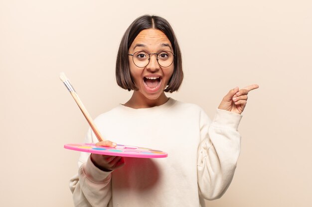 Young hispanic woman laughing, looking happy, positive and surprised, realizing a great idea pointing to lateral copy space