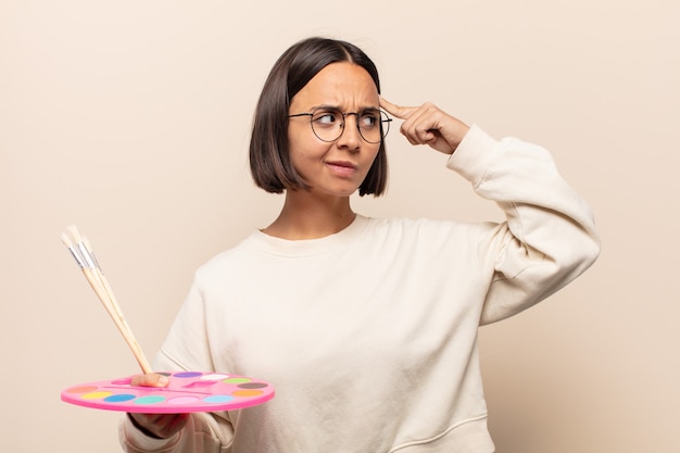 Young hispanic woman looking happy and smiling with hand on chin, wondering or asking a question, comparing options