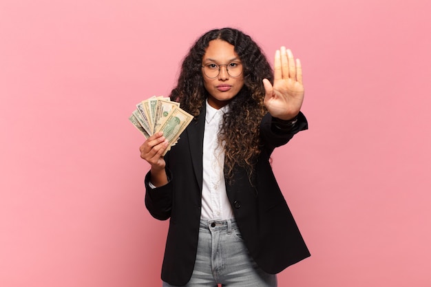 Young hispanic woman looking serious, stern, displeased and angry showing open palm making stop gesture. dollar banknotes concept