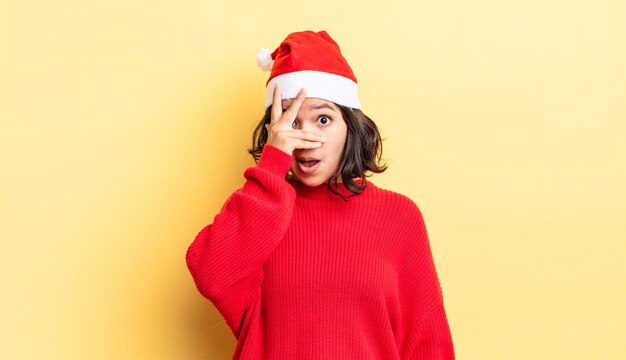 Young hispanic woman looking shocked, scared or terrified, covering face with hand. christmas concept