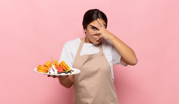 Young hispanic woman looking shocked, scared or terrified, covering face with hand and peeking between fingers