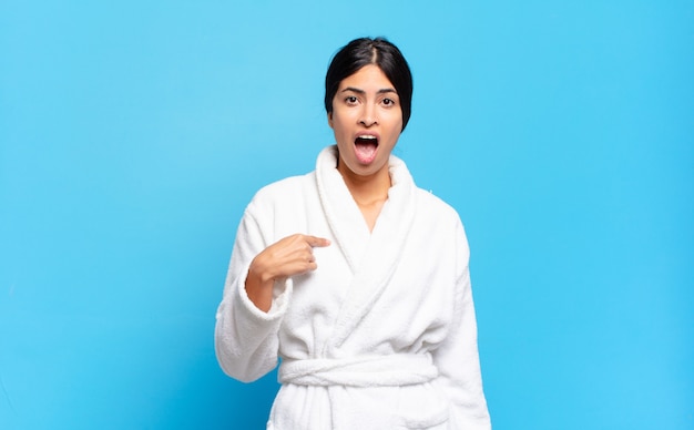 Young hispanic woman looking shocked and surprised with mouth wide open, pointing to self. bathrobe concept