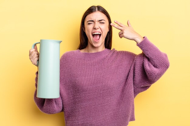 Young hispanic woman looking unhappy and stressed, suicide gesture making gun sign. coffee thermos concept