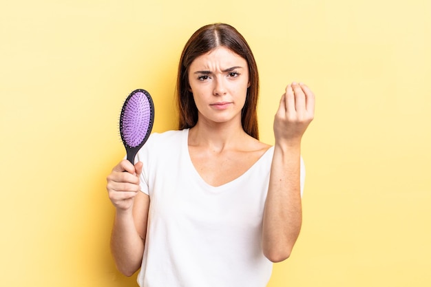 Young hispanic woman making capice or money gesture, telling you to pay. hair brush concept