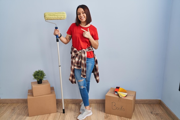 Young hispanic woman painting home walls with paint roller cheerful with a smile on face pointing with hand and finger up to the side with happy and natural expression