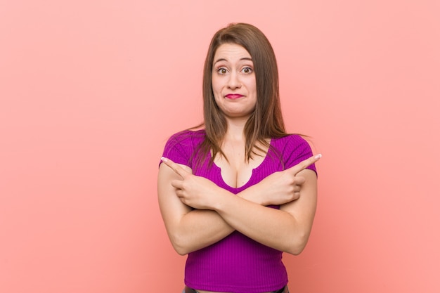 Young hispanic woman on pink wall points sideways, is trying to choose between two options.