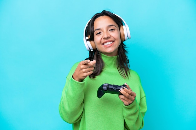 Young hispanic woman playing with a video game controller isolated on blue background pointing front with happy expression