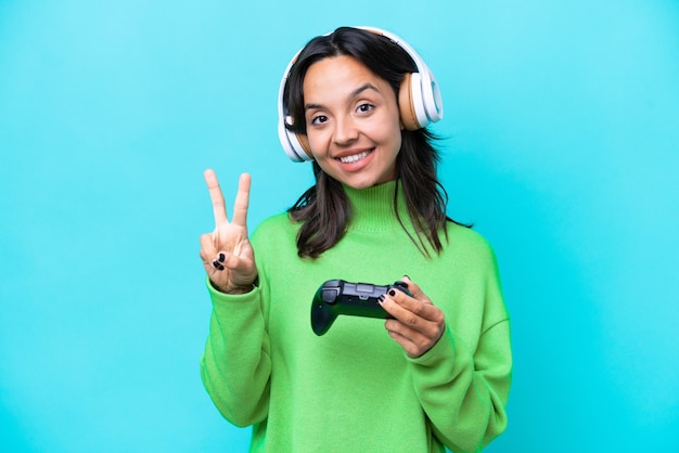 Young hispanic woman playing with a video game controller isolated on blue background smiling and showing victory sign