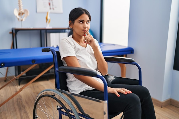 Young hispanic woman sitting on wheelchair at physiotherapy clinic thinking concentrated about doubt with finger on chin and looking up wondering