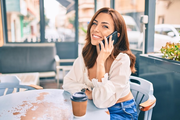 Young hispanic woman smiling happy using smartphone sitting at the coffee shop terrace