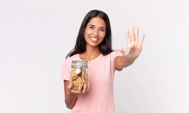 Young hispanic woman smiling and looking friendly, showing number five and holding a cookies glass bottle