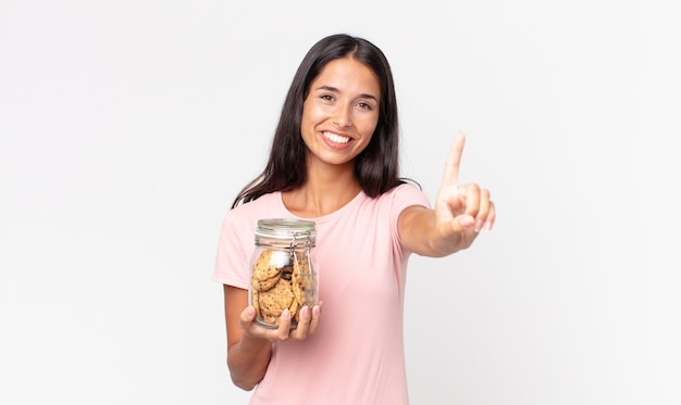 Young hispanic woman smiling and looking friendly, showing number one and holding a cookies glass bottle