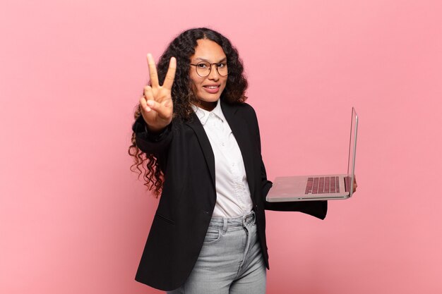 Young hispanic woman smiling and looking happy, carefree and positive, gesturing victory or peace with one hand. laptop concept